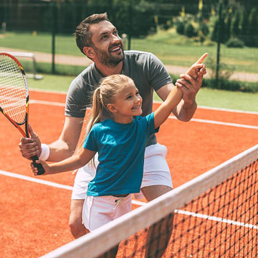 Cheerful father in sports clothing teaching his daughter to play tennis while both standing on tennis court